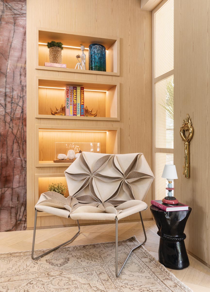 A corner vignette in the living room features a curious AntiBodi chair by Patricia Urquiola with sculpture-like upholstery that never fails to inspire conversation. Display shelves lit from within showcase the homeowners’ meaningful collectibles, composing an eye-catching still-life. A black lacquered accent table highlights a bronze sculptural wall hanging above.