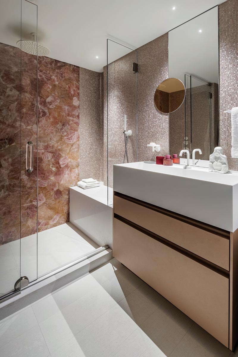 The powder room’s exotic aesthetic includes a natural rose quartz shower wall and pink metal mosaic tiles cladding the remaining three walls. A metal vanity cabinet by Ornare and a circular mirror echo the pink palette, while a white countertop, fixtures, and flooring balance the color scheme.