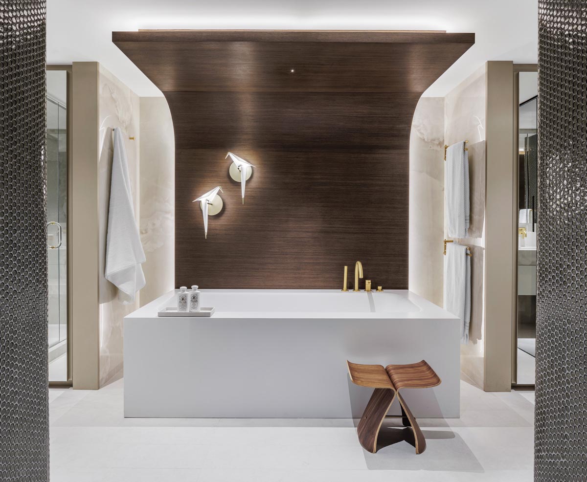 A water-resistant wood panel ceiling and accent wall bring an element of warmth to the all-white primary bathroom. A massive custom bathtub centers the space, and matching vanities flanking the entrance create a sense of symmetry. Robern medicine cabinets and full-height closets at each side of the tub provide generous storage.