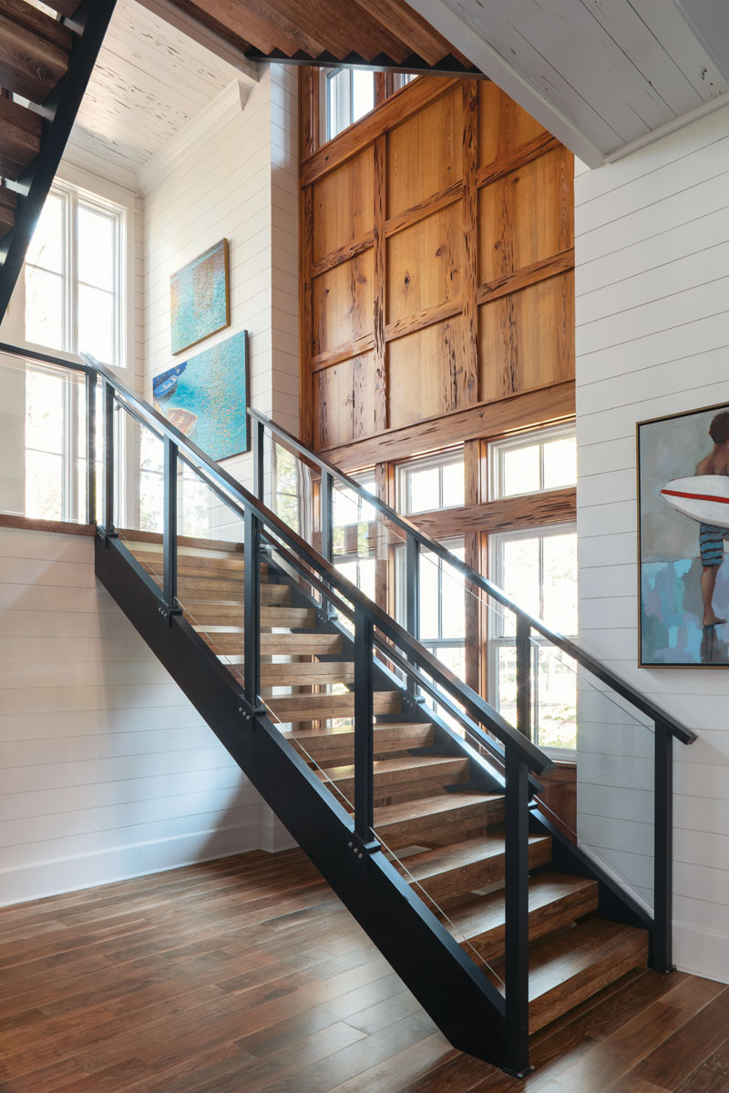 The three-story floating staircase features glass balusters for a clean, open aesthetic. Downlit hand railings lend a glow at night. Windows stretching up all three stories flood the space with natural light, and a pecky cypress-paneled wall provides texture..