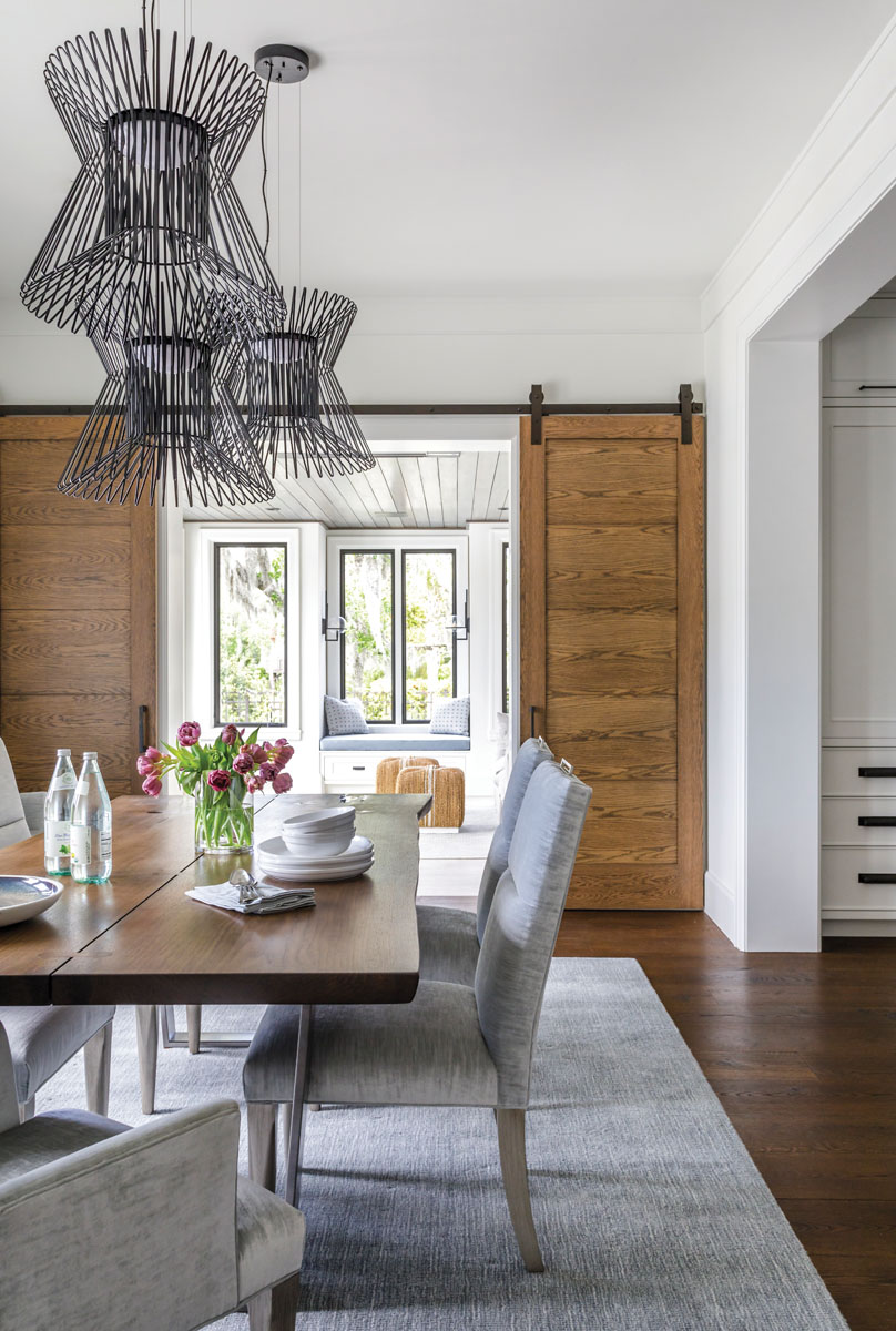 Stationed beneath a staggered grouping of modern black pendants, a live edge dining room table from Italy sets the scene for everything from casual meals to intimate dinner parties. Sliding barn doors in figured oak open to the solarium where the children can play.