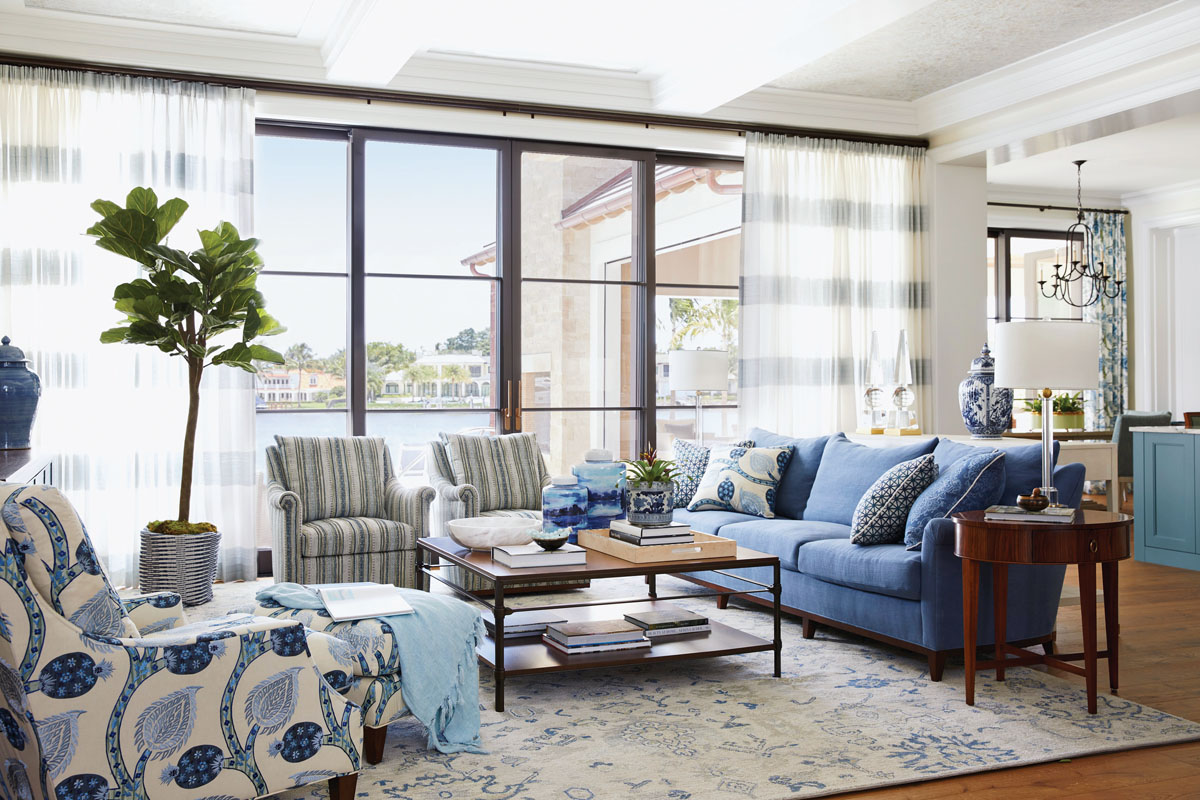 The home was designed to capitalize on the water views with floor-to-ceiling windows. In this great room tableau, the seating area riffs a blue and gray color color palette with fabrics from Schumacher and Cowtan and Tout. The ceiling here is finished with layers of capiz shell covered in a light layer of Venetian plaster, which gives the room its pearlescent glow.