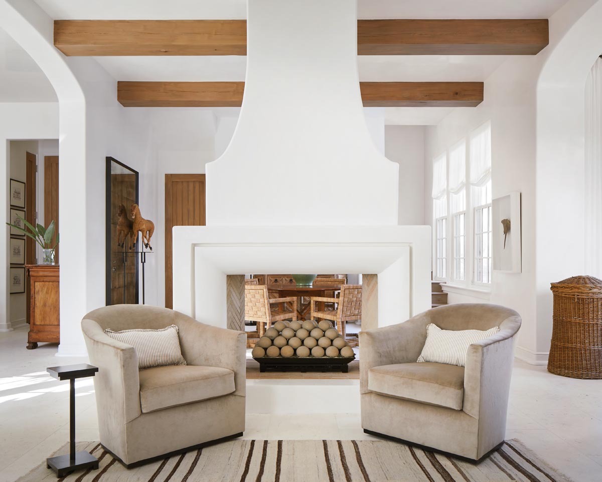 Separated by a central, double-sided plaster-formed fireplace, the living room’s seating area connects visually to the dining room. A striped rug from Scott Antiques anchors a pair of cozy club chairs from Lee Industries and a custom pedestal drink table designed by David Frazier.