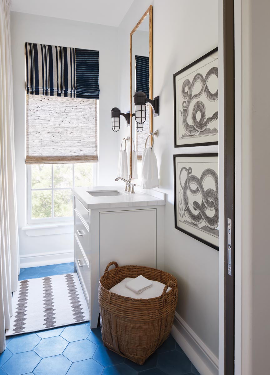 A guest bathroom often reserved for the kids emits a playful vibe with its bright blue hexagon-tile floor. Visually striking contrasts include the white marble-topped vanity paired with black sconces and a black-and-white striped window shade.