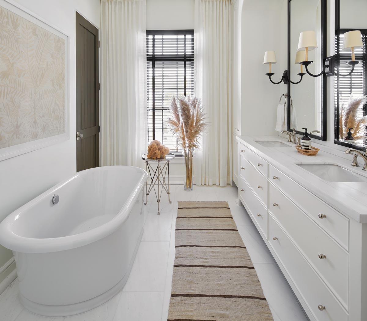 White cabinets, white-veined marble, and a freestanding Kohler tub give an airy spa-like ambiance to the primary bathroom. A sheer unlined drapery from Fabricut brings an ethereal touch to the contrasting black Venetian blinds. An antique nickel Directoire table and striped antique kilim rug ensure an eclectic layered look.
