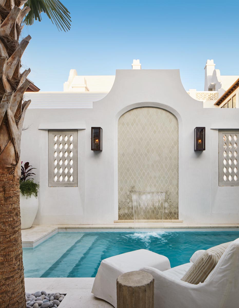 The all-white stucco, white concrete roof, and blue sky provide a brilliant backdrop for the plunge pool and towering palm tree. Flanked by Bevolo sconces from Architectural Heritage, the fountain tile’s hand-formed arabesque design lends itself to the Mediterranean feel that pervades Alys Beach.