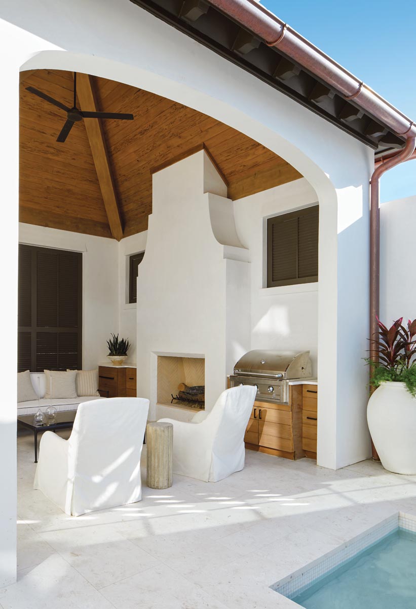 The loggia features a soaring wood-slatted ceiling, a magnificent fireplace wall, and a grilling station. Crisp white slipcovered slipper chairs from Lee Industries and side tables from Elegant Earth complement a generously scaled sofa from Amalfi Living.