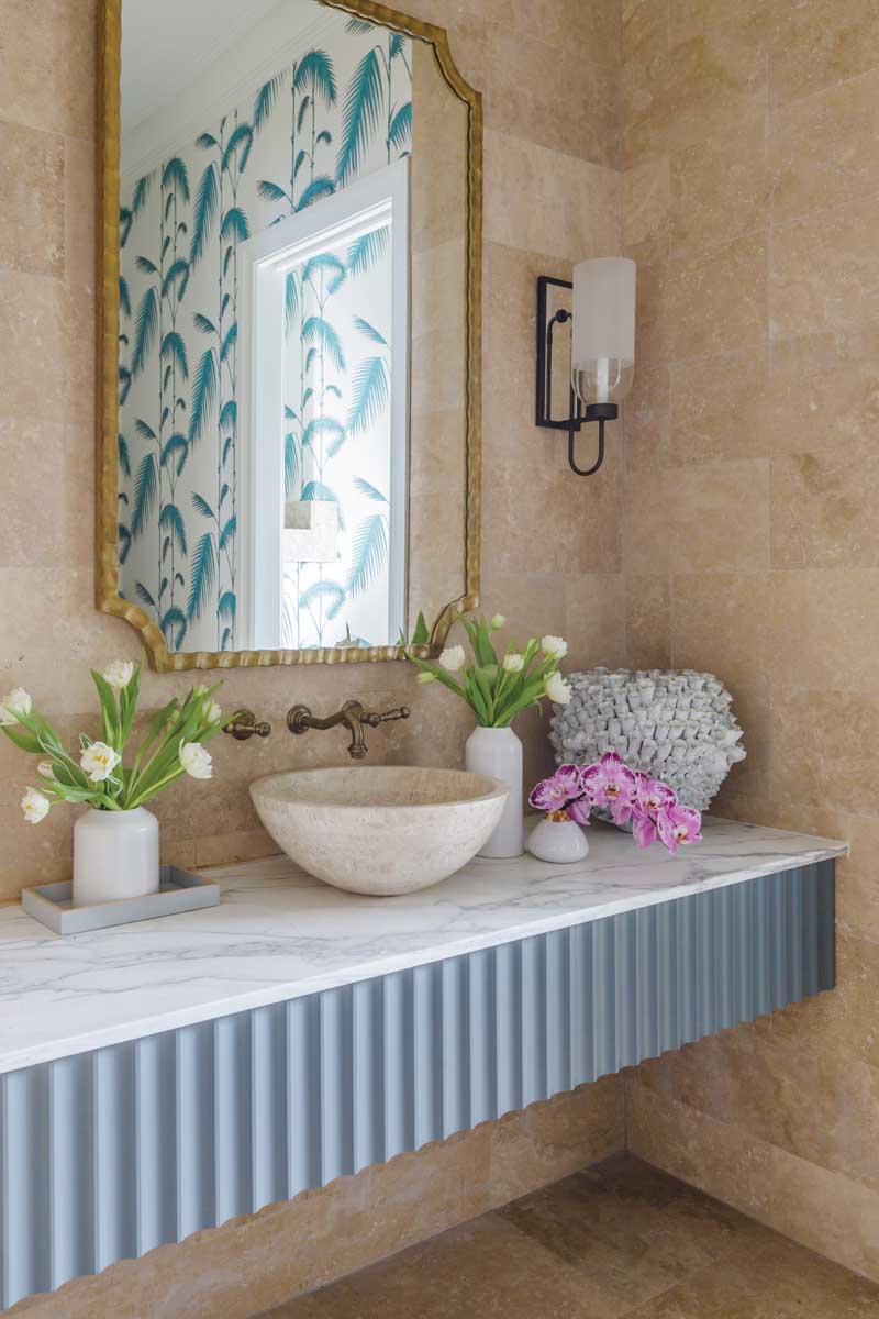 The floors and walls in the formal powder room are covered in travertine. The bright Cole & Son palm leaf wallpaper reflected in the mirror was chosen because it offers a more whimsical take on the traditional palm leaf that has been a staple in Florida design for ages.