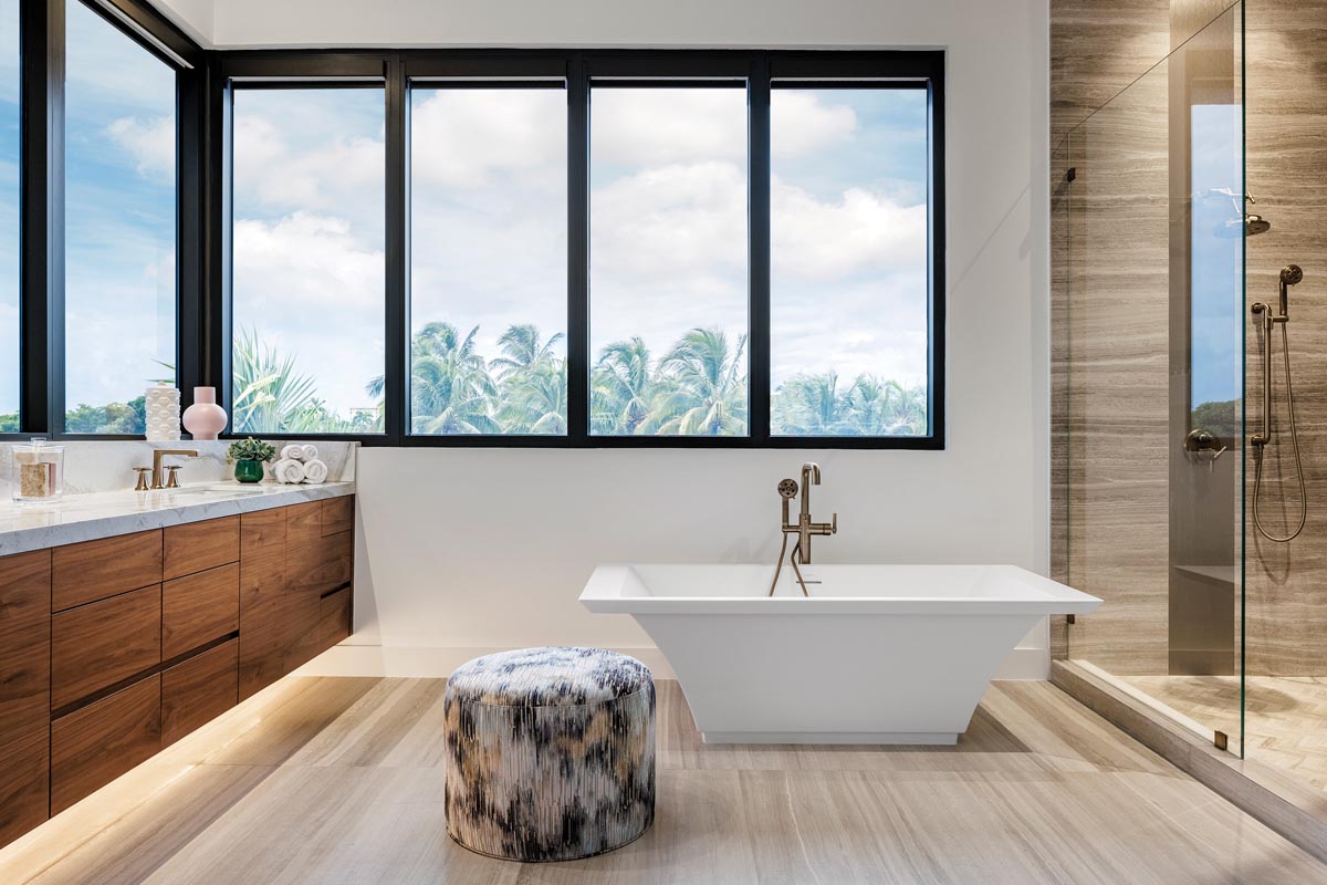 The primary bathroom lends an organic vibe with limestone walls from Opustone and Vetrite glass paneling in the shower from Ceramic Matrix. Bathed in natural light, the space emanates a spa-like aura with an elegant Kallista soaking tub accented with a plush ottoman.