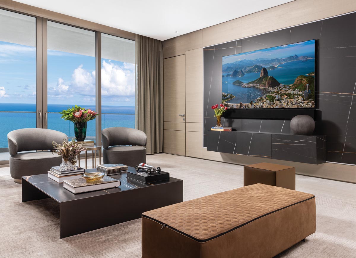 The living area captures stunning ocean views, contributing to the sleek, serene aesthetic of this Sunny Isles Beach dwelling. Artefacto armchairs combine with an elegant, clean-lined Minotti ottoman, leather cube, and cocktail table to articulate an intimate seating group. MC Studio designed the dynamic black porcelain wall feature.