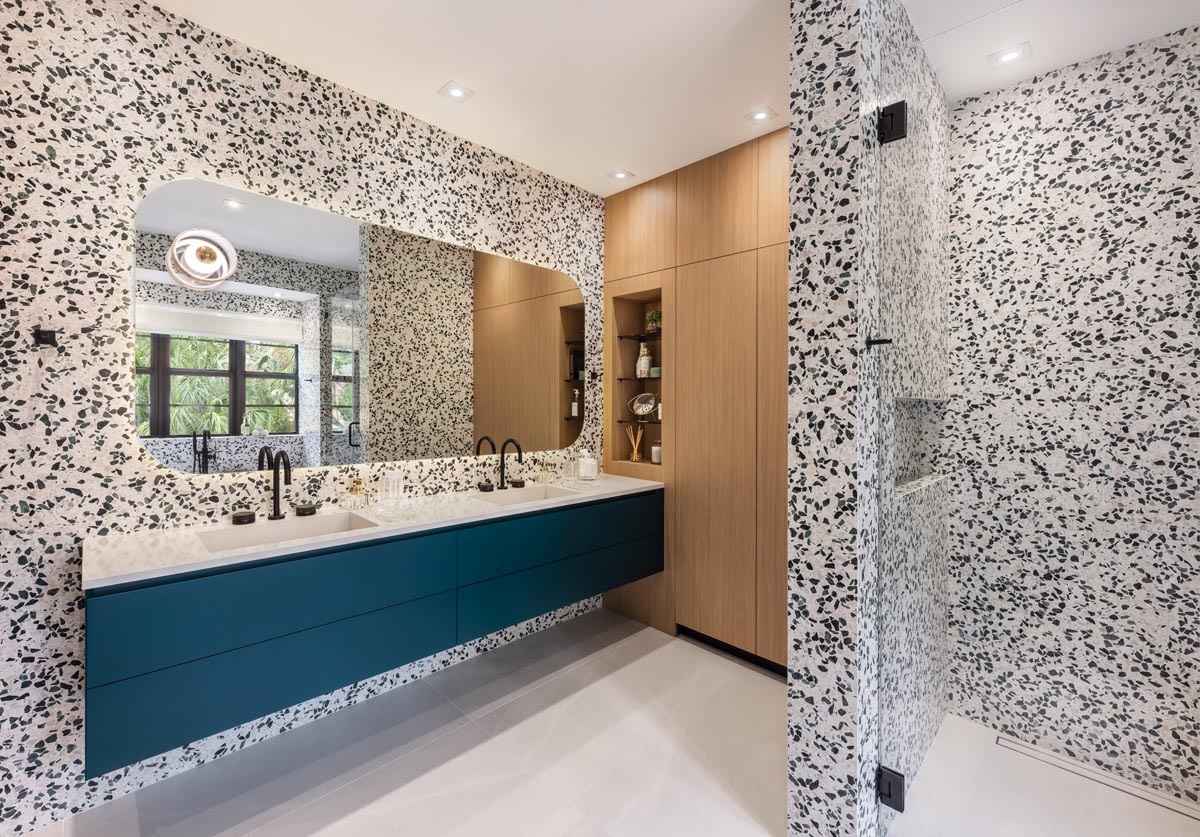 The primary bathroom emits a playful look with its speckled Tivoli terrazzo walls and shower area. Vibrant blue Miacucina vanity cabinets introduce a pop of color, and a graceful, rounded mirror highlights the lush landscape outside. Built-in shelving and storage reiterate the clean, contemporary feel that pervades the home.